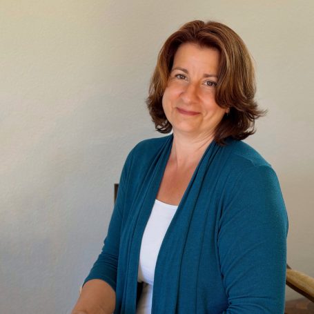 Lächelnde Frau mit braunen Haaren, trägt einen blauen Cardigan und sitzt entspannt.